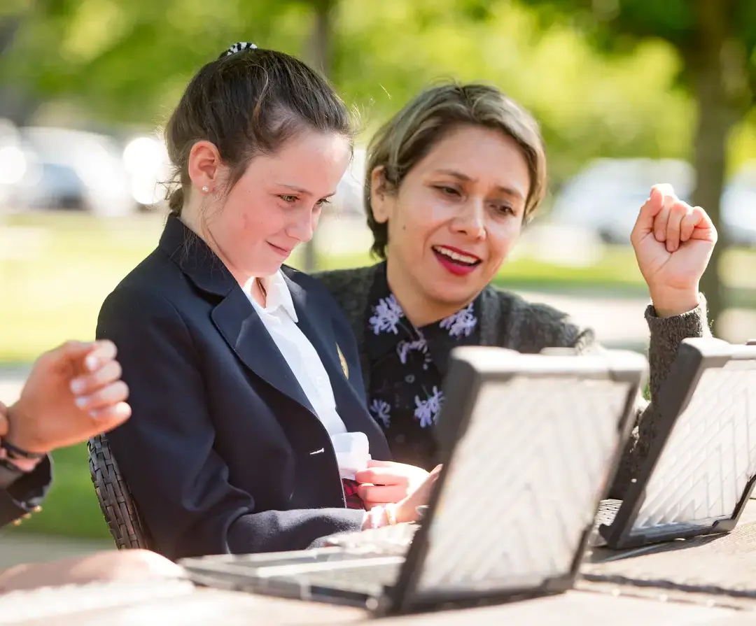King School teacher sitting outside and helping a student with classwork on a laptop.